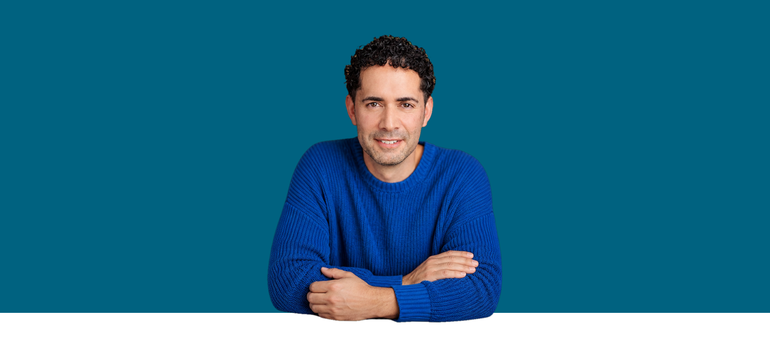 Jon Hunter leaning forward with folded arms, wearing a blue knit sweater against a petrol-blue background.