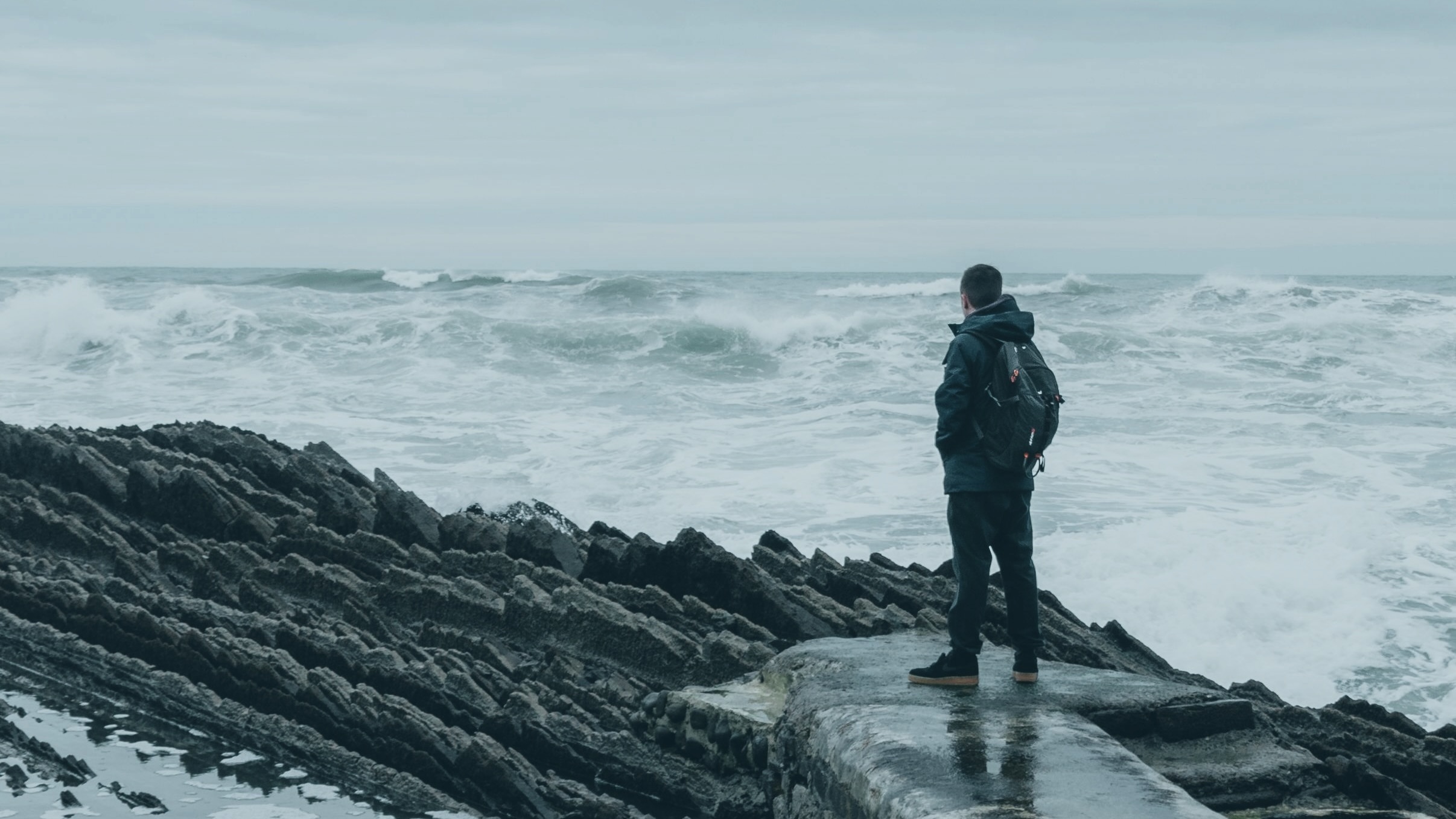 A man stands on dark rocks, facing a wild grey sea
